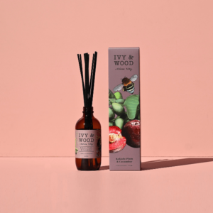 IVY & WOOD 卡卡杜李與青瓜室內擴香瓶與外盒單品照。在乾淨的純莫蘭迪粉色背景下，左側為琥珀色玻璃擴香瓶搭配 200ml 容量與黑色擴香竹，右側為包裝外盒，盒身印有細緻的蜜蜂與綠色植物果實插畫，呈現簡約現代的精品電商商品照風格。