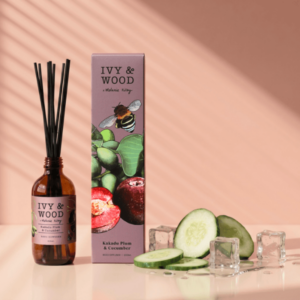 IVY & WOOD 卡卡杜李與青瓜室內擴香瓶夏日去悶熱情境照。低飽和暖粉色背景與斜射的百葉窗光影下，左側並列琥珀玻璃香氛瓶與插畫外盒，右下角檯面擺放新鮮切開的小黃瓜切片與晶瑩剔透的冰塊水滴折射，凸顯清爽降溫的草本水潤感。